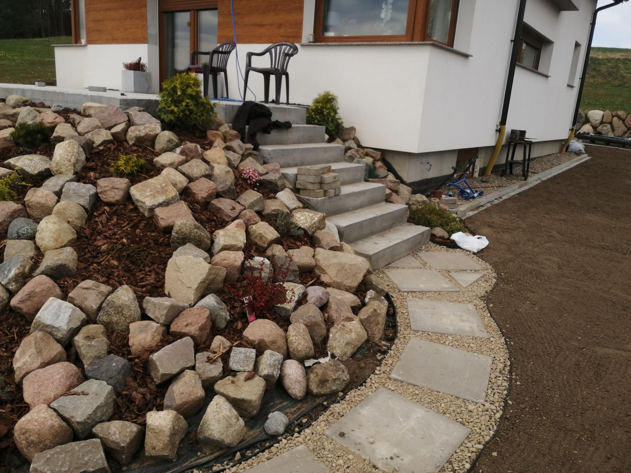 Ukończone schody wejściowe do domu z betonowych płyt, otoczone kamieniami, korą i roślinami, z wytyczoną ścieżką z płyt chodnikowych i żwiru.