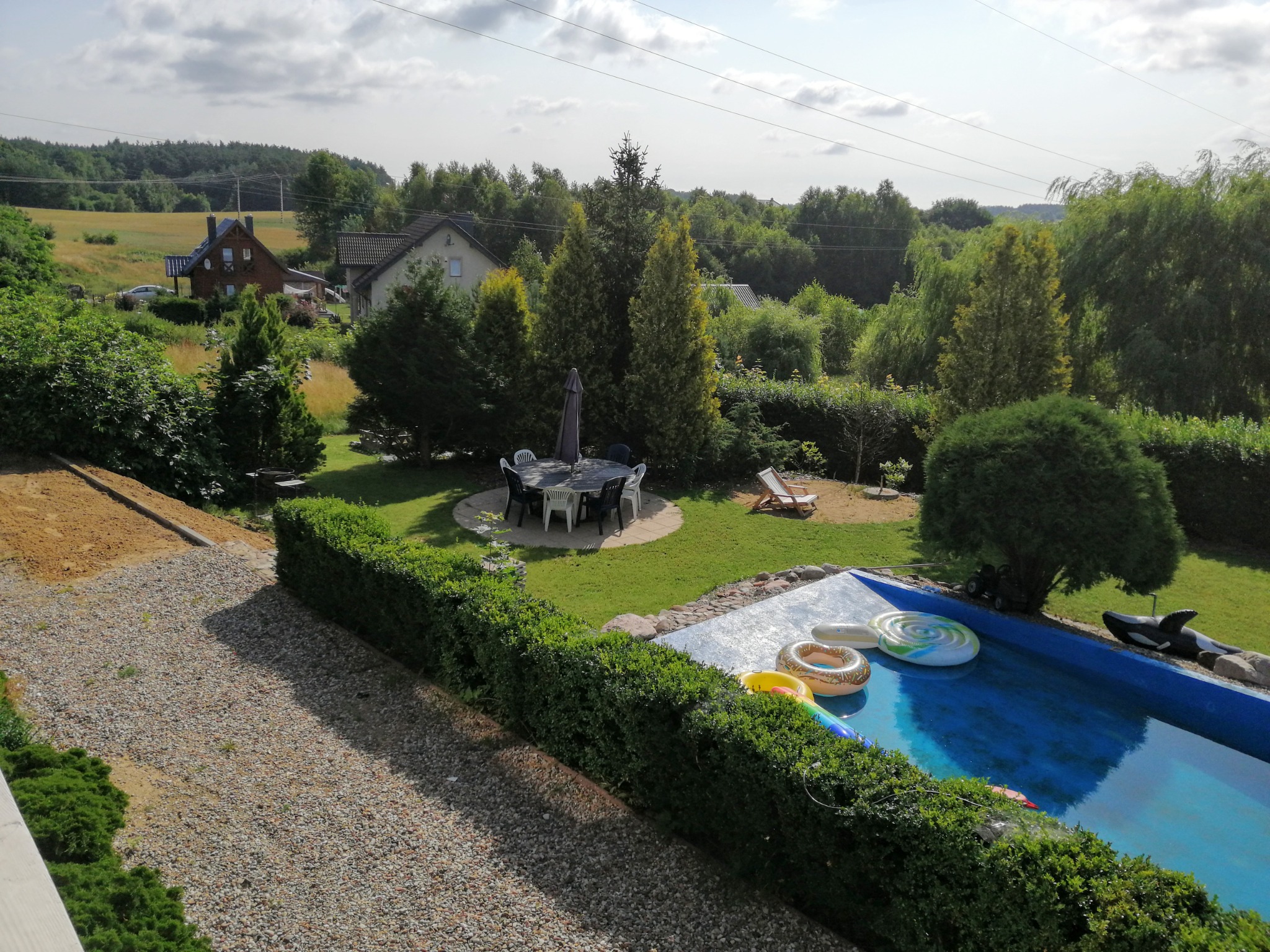 Widok z góry na zadbany ogród z basenem, otoczony żywopłotem i drzewami, z widocznym zestawem mebli ogrodowych i domami w tle.