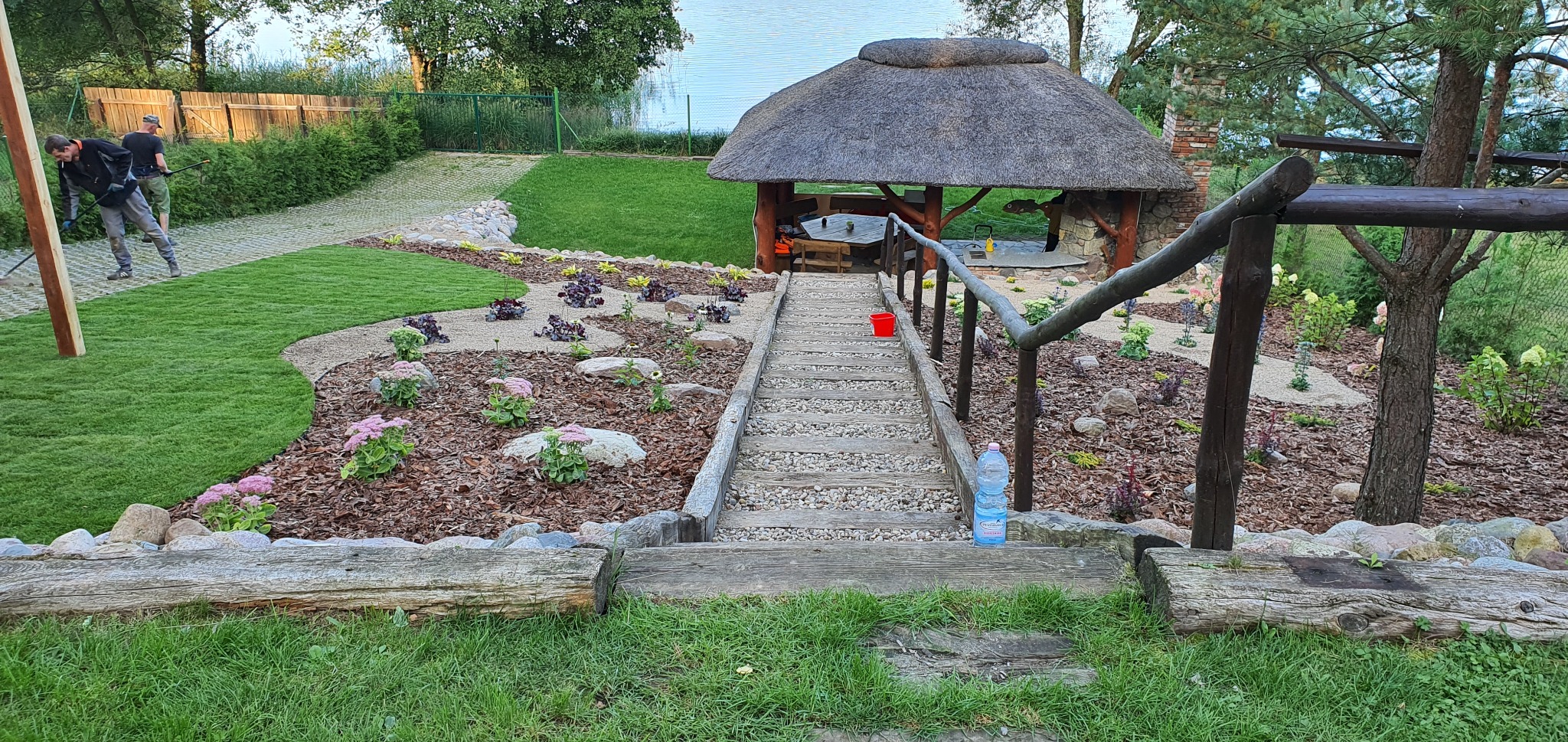 Ogród z drewnianą altaną krytą strzechą, ścieżką z drewnianych stopni i kamieni, rabatami z korą, kwiatami i kamieniami. Widoczni pracownicy w tle.