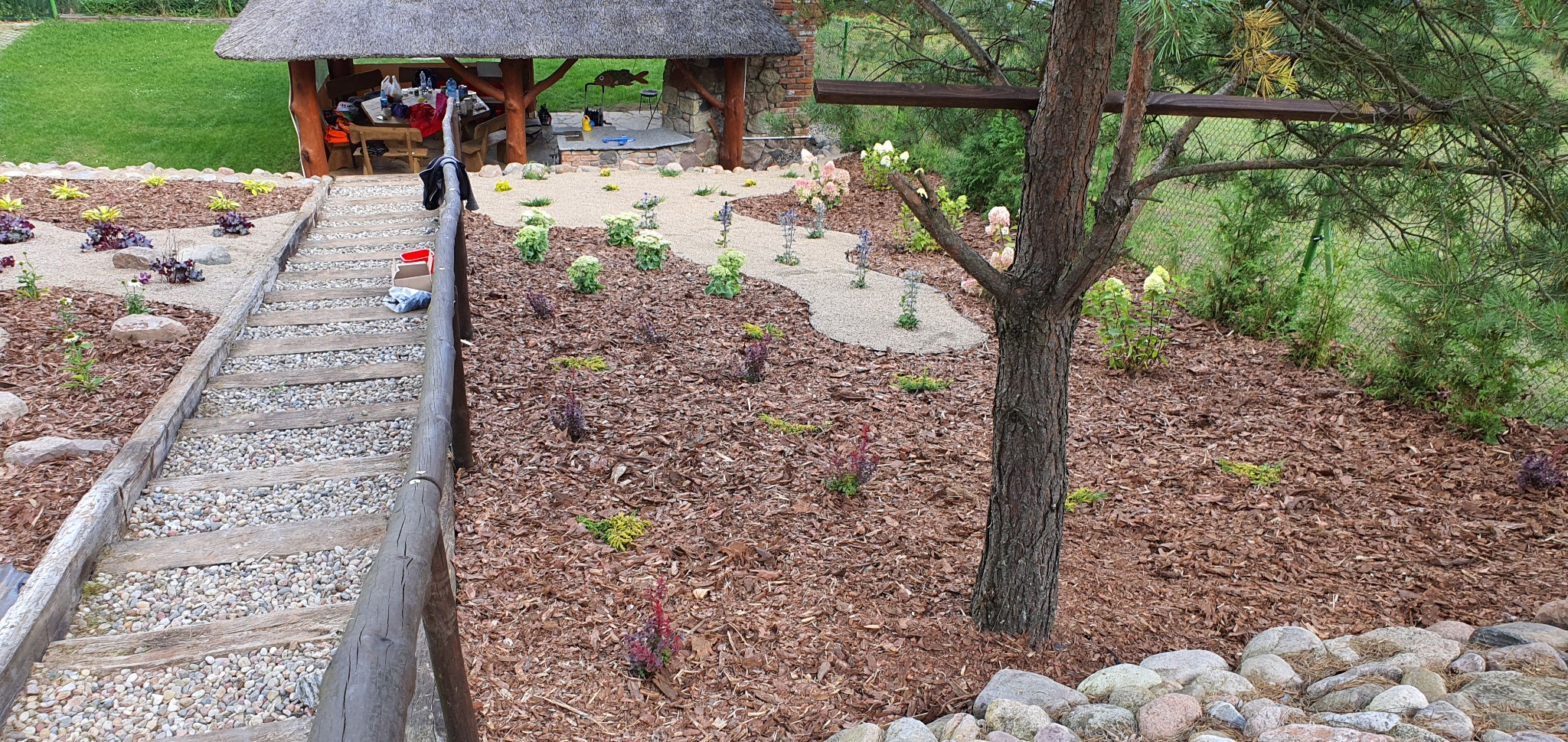 Ogród z drewnianymi schodami wypełnionymi żwirem, rabatami z korą, ścieżką z jasnego żwiru i altaną z trzcinowym dachem w tle.