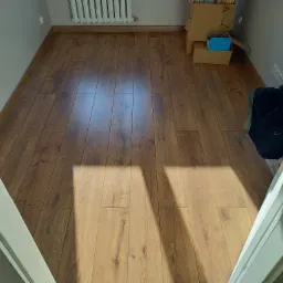 Ułożone panele podłogowe w odcieniach brązu, widoczne kartony i grzejnik w tle, fragment futryny drzwiowej w kadrze.