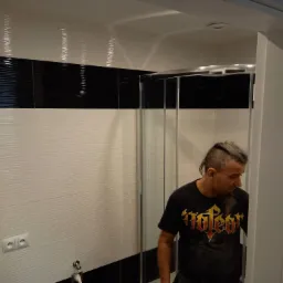 Montaż kabiny prysznicowej w łazience z biało-czarnymi płytkami; widoczny pracownik w czarnym T-shircie z logo zespołu metalowego.