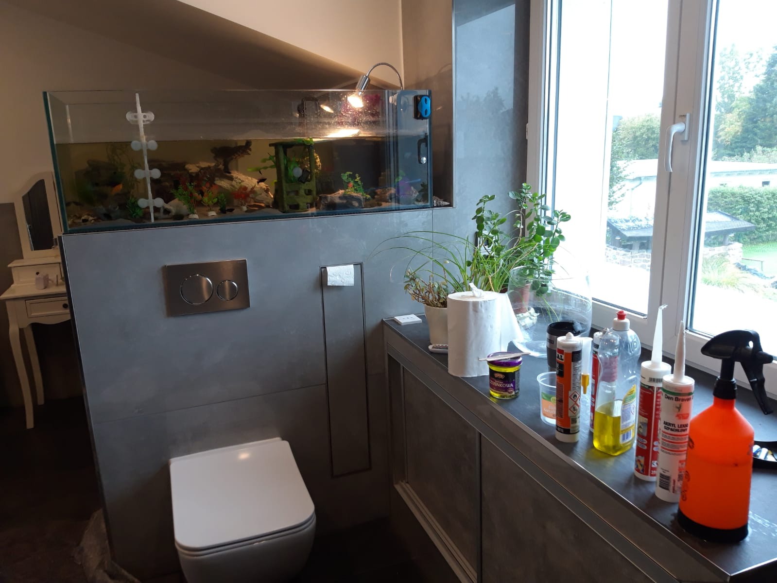 Nowoczesna łazienka z akwarium wbudowanym w ścianę, widoczne materiały budowlane i rośliny na blacie przy oknie, szara faktura ściany i biała toaleta.