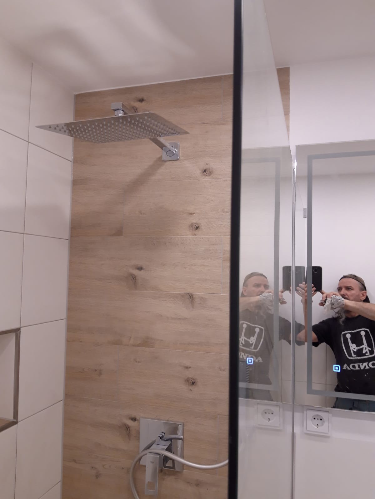 Nowoczesna kabina prysznicowa z deszczownicą, wykończona płytkami imitującymi drewno, z odbiciem osoby robiącej zdjęcie w lustrze.