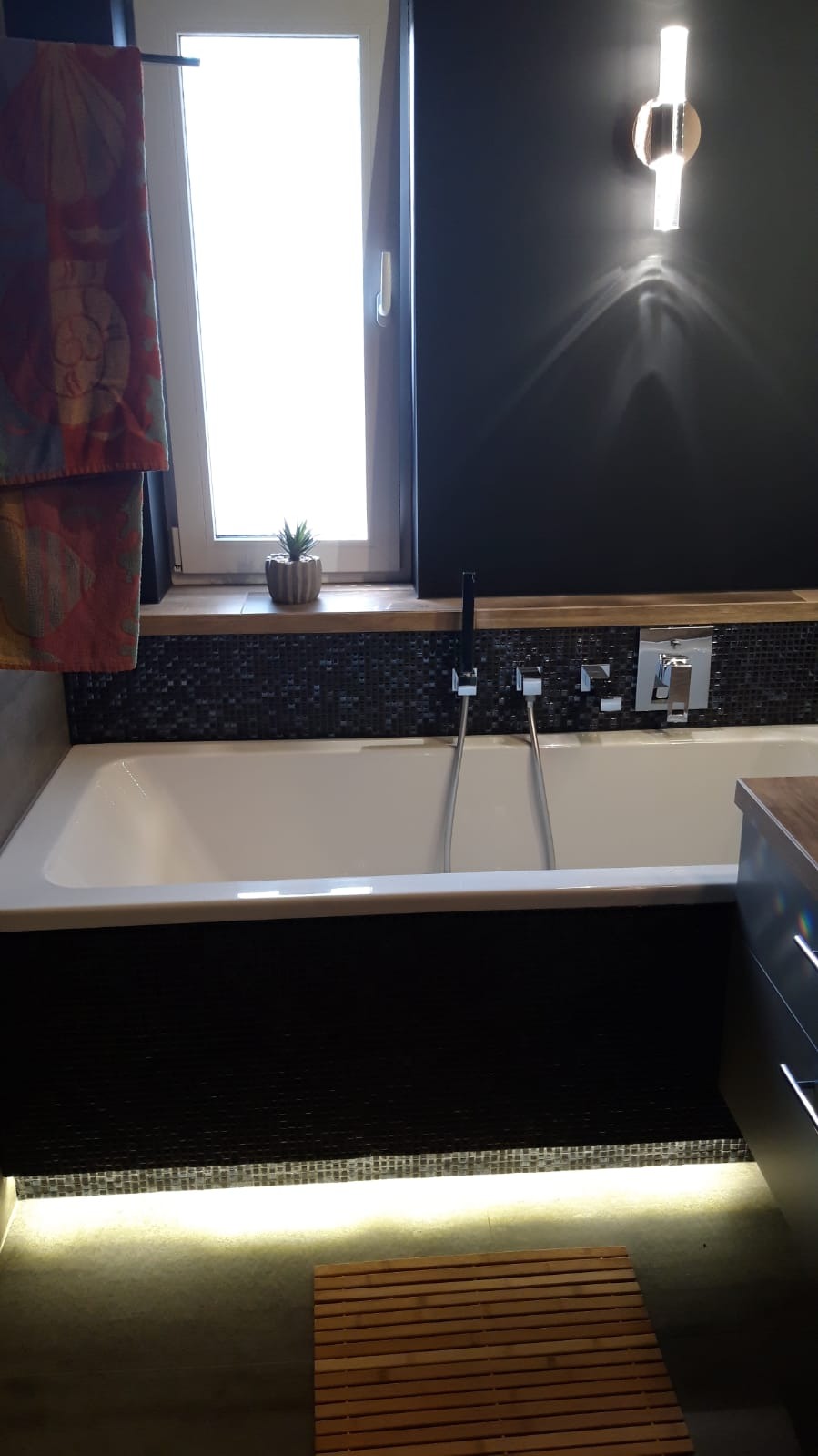 Nowoczesna łazienka z wanną obudowaną czarną mozaiką, podświetleniem LED i drewnianym podestem, okno z rośliną doniczkową i designerski kinkiet na czarnej ścianie.