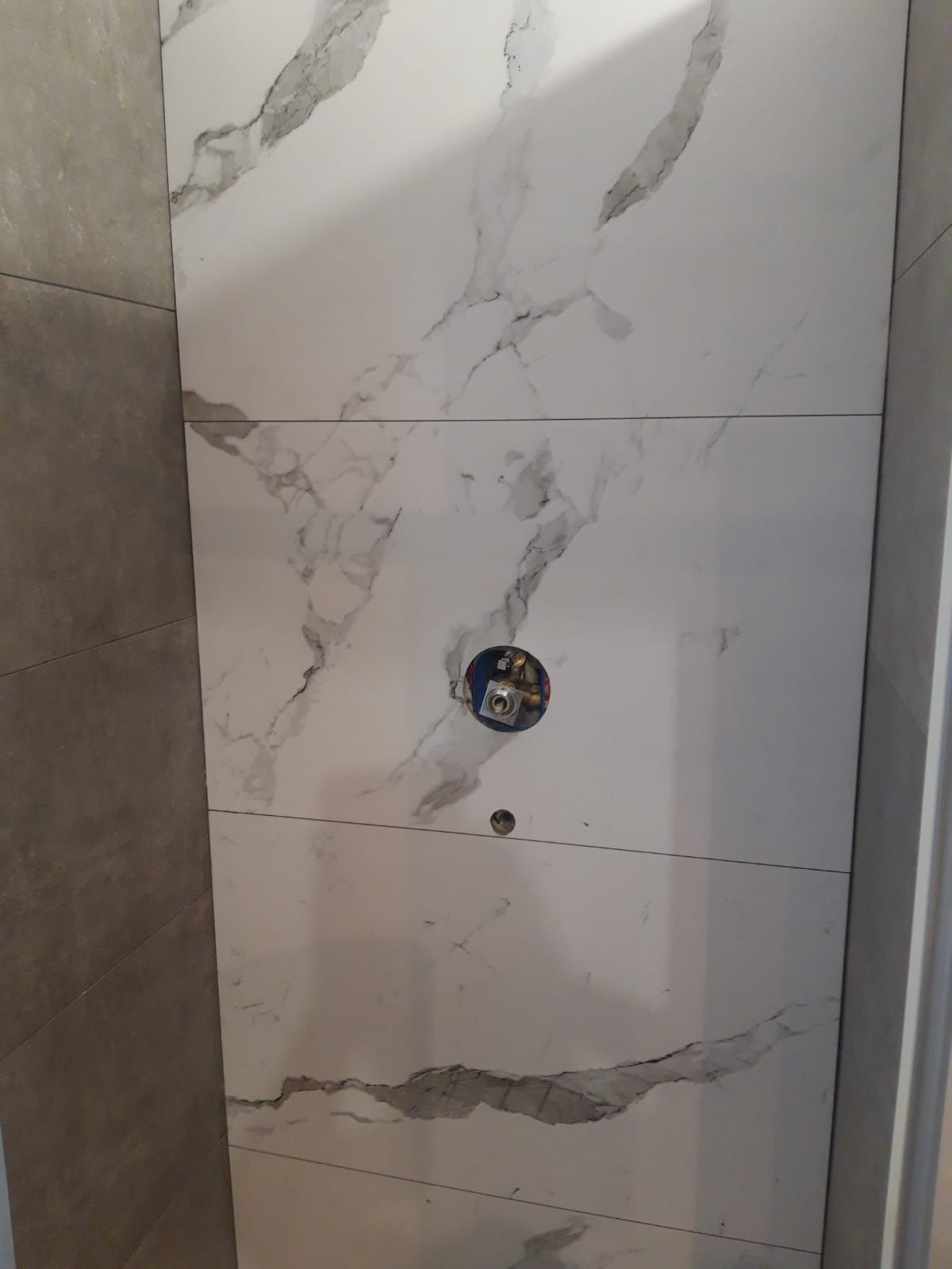 Ściana łazienki wyłożona białymi płytkami imitującymi marmur z widocznym otworem na baterię i odpływ w trakcie prac wykończeniowych.