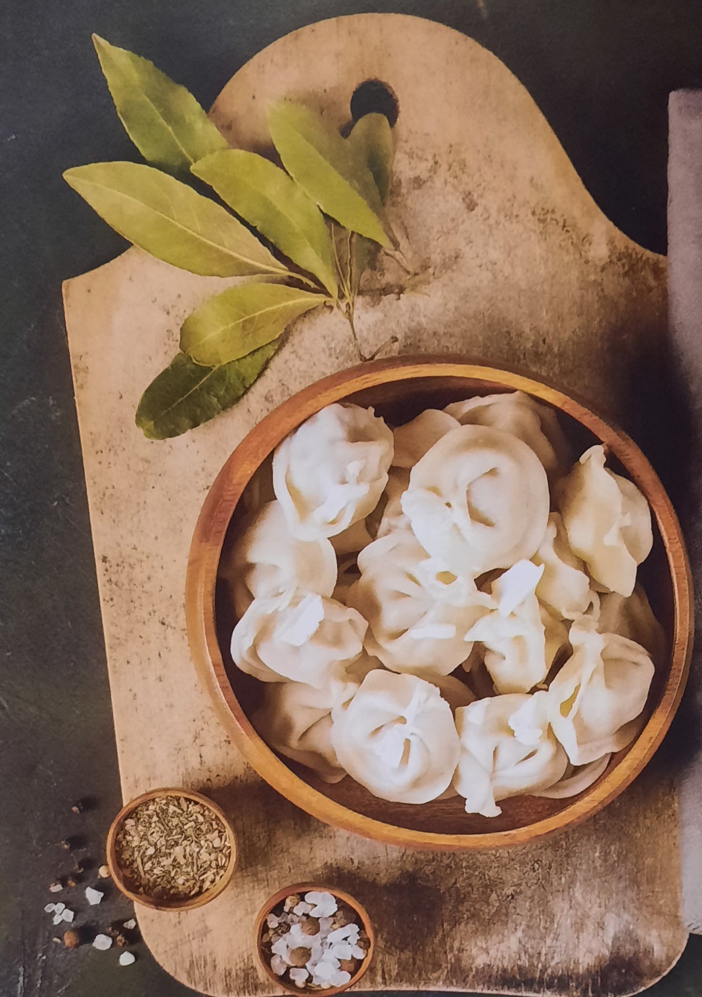 Ręcznie robione pierogi w drewnianej misce, udekorowane gałązką laurową, z przyprawami w małych miseczkach na desce do krojenia.