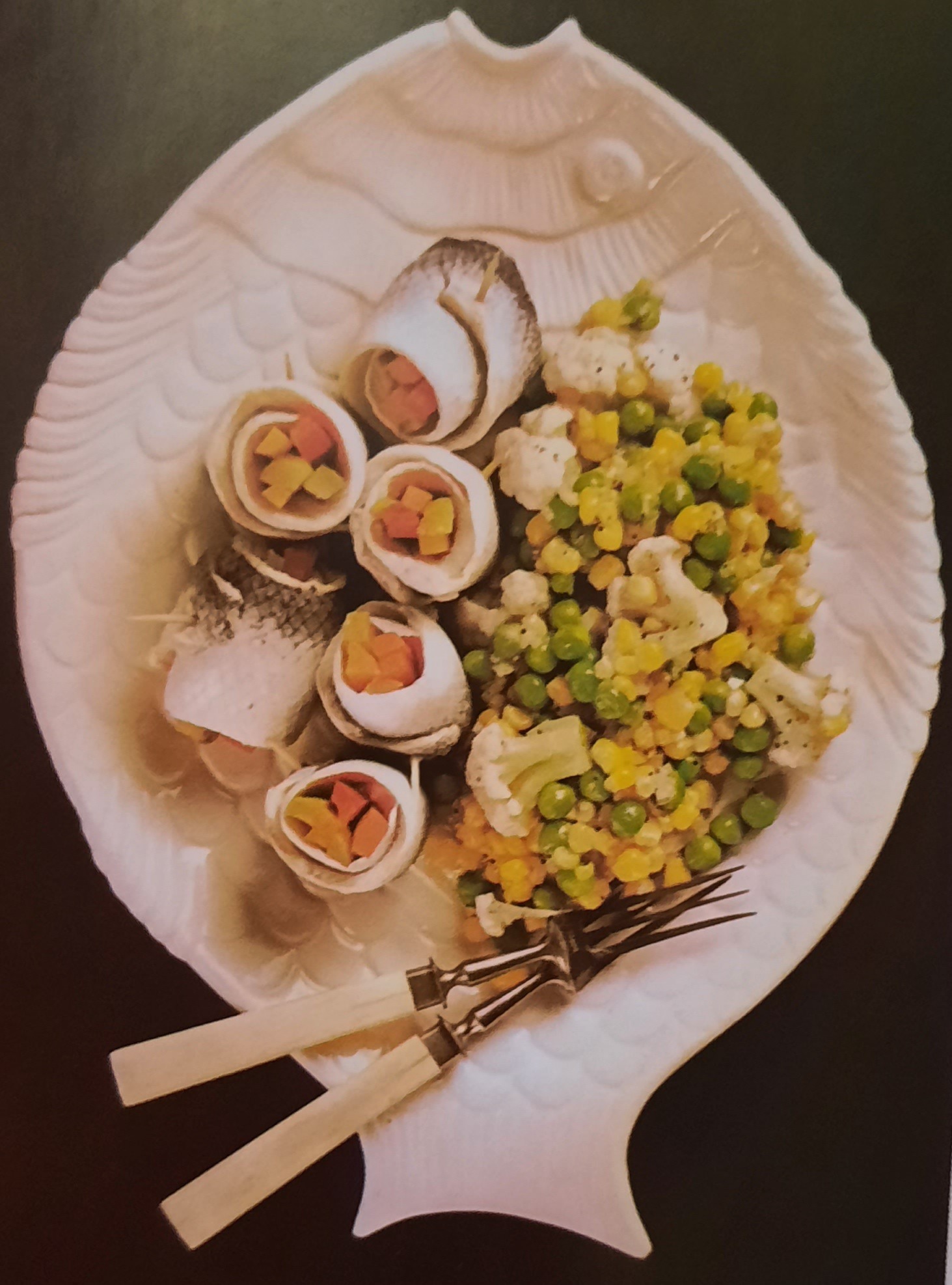 Roladki rybne nadziewane warzywami na talerzu w kształcie ryby, obok mieszanka warzyw z groszkiem i kalafiorem, oraz widelec z jasną rączką.