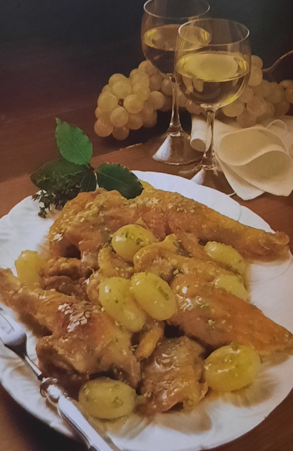 Wykwintne danie z pieczonego mięsa z białymi winogronami na białym talerzu, widelcem obok, w tle dwa kieliszki białego wina i kiść winogron.