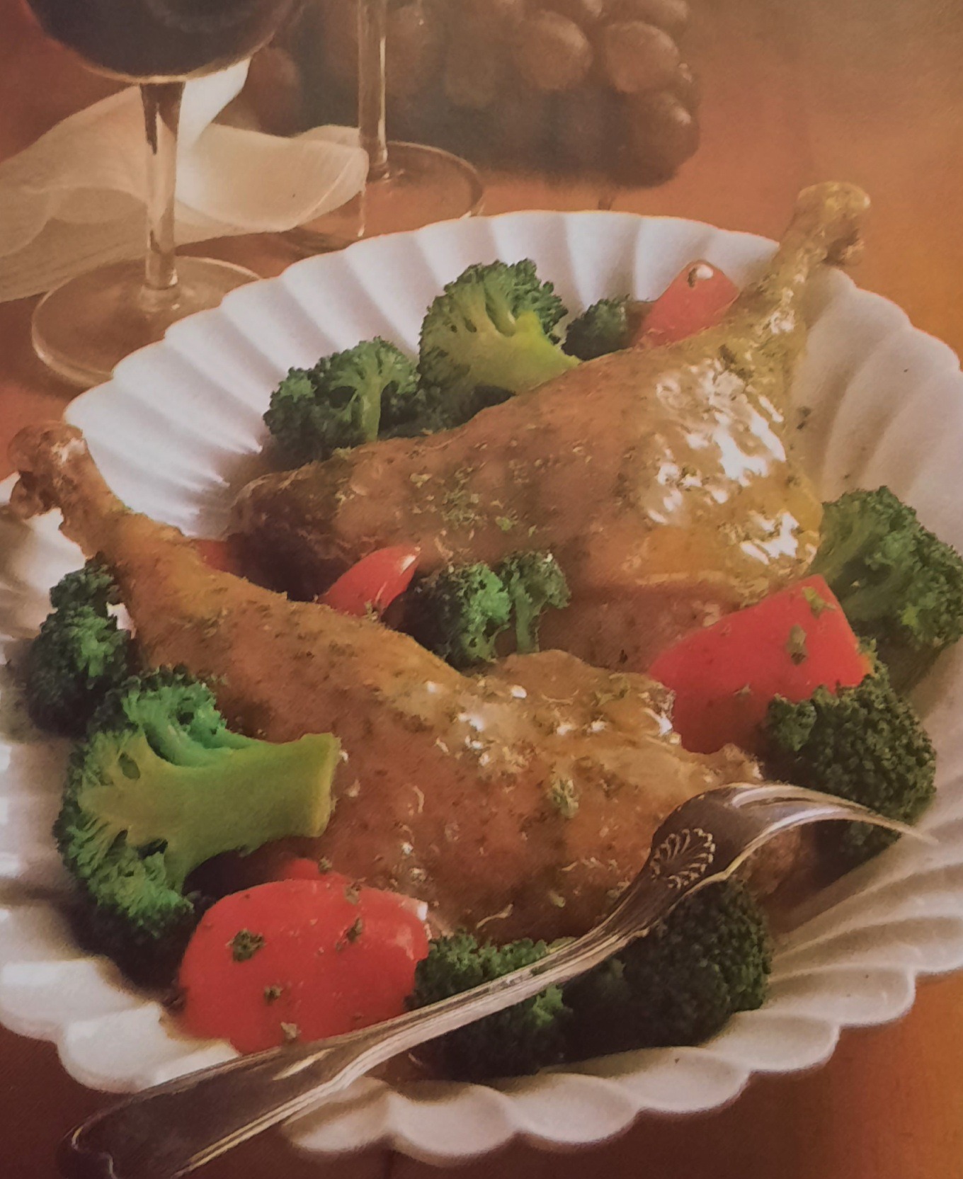 Dwa udka pieczonego kurczaka polane sosem, podane na białym talerzu z brokułami i kawałkami pomidora, obok leży srebrny widelec, w tle kieliszki z czerwonym winem i winogrona.