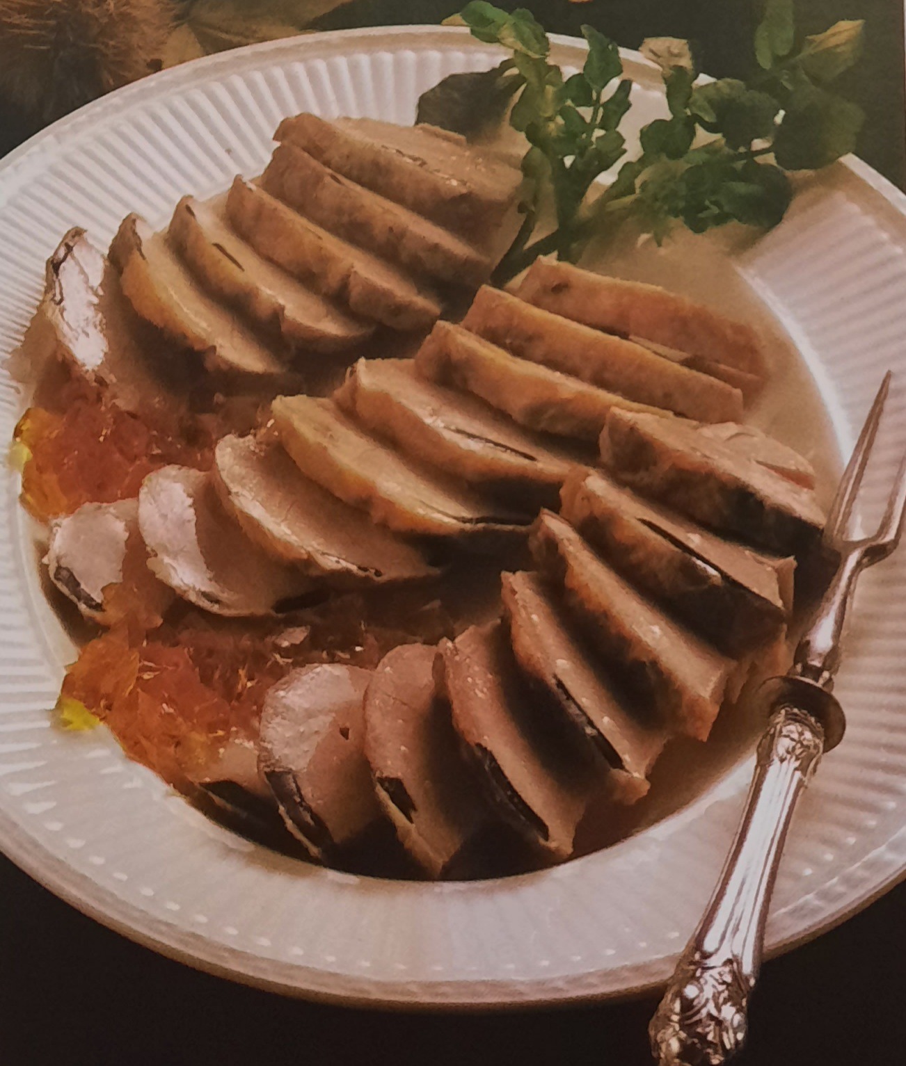 Plasterki pieczonego mięsa ułożone na białym talerzu z dekoracją z sałaty i pomarańczowej galaretki, obok srebrny widelec.