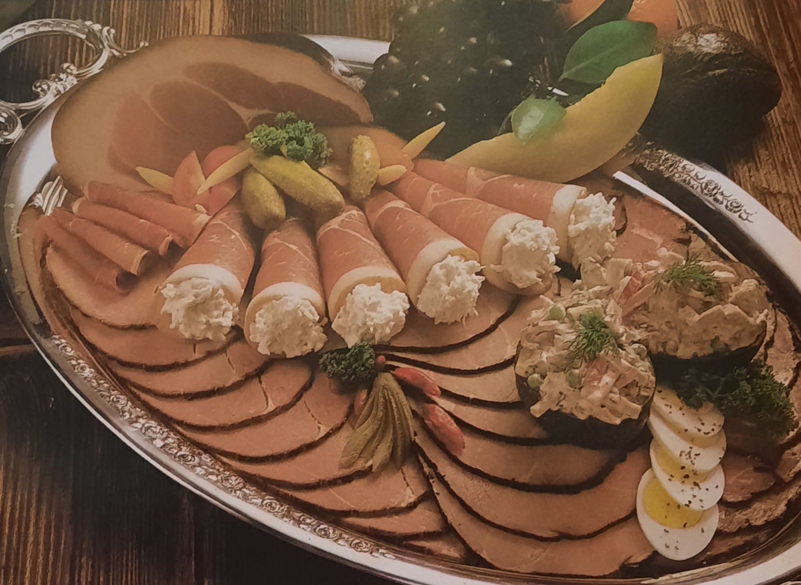 Srebrna taca z wędlinami, roladkami szynkowymi z białym farszem, sałatką jarzynową, jajkiem na twardo i owocami na drewnianym stole.