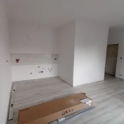 Pusta, częściowo wykończona kuchnia w trakcie remontu; widoczne przygotowane ściany pod malowanie, instalacja hydrauliczna, podłoga panelowa i karton z materiałami budowlanymi.