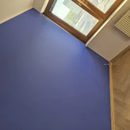 Świeżo pomalowana na ciemny fiolet ściana z drewnianym oknem dachowym, widok na miasto i park, parkiet ułożony w jodełkę, kaloryfer na białej ścianie.