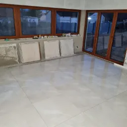 Jasne, błyszczące płytki podłogowe w nowo wykończonym pomieszczeniu z dużymi oknami, widoczne płytki przygotowane do położenia przy ścianie.