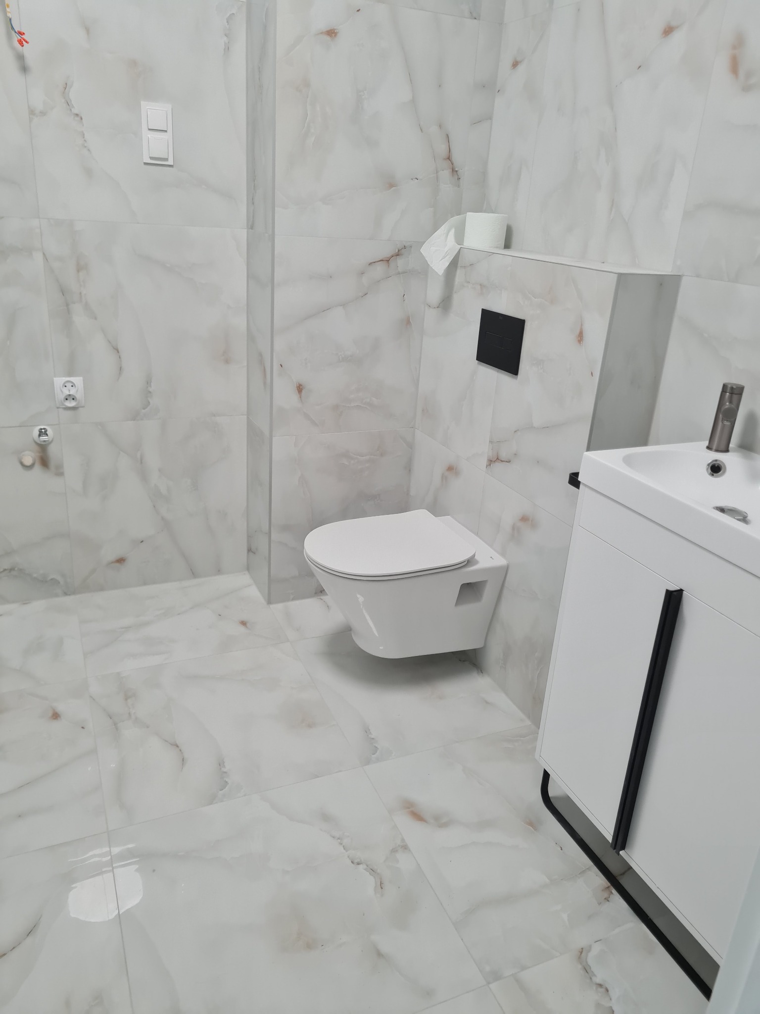 Wykończona łazienka z białymi marmurowymi płytkami, podwieszaną toaletą i białą szafką pod umywalką z czarnymi akcentami.