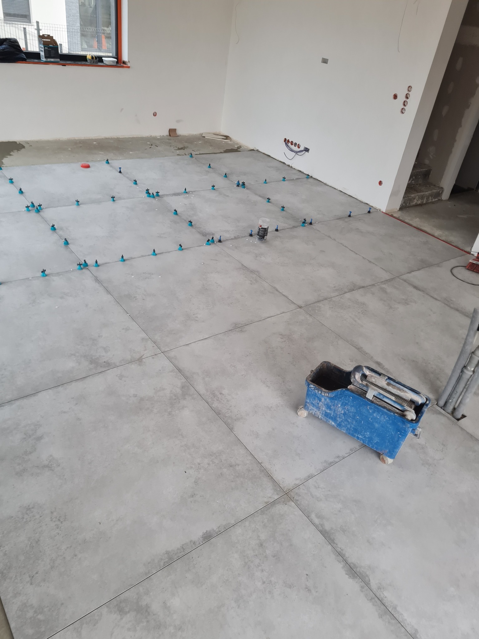 Szare płytki podłogowe w trakcie układania, użyte niebieskie klipsy poziomujące, w tle widoczny pojemnik na zaprawę i fragment schodów.