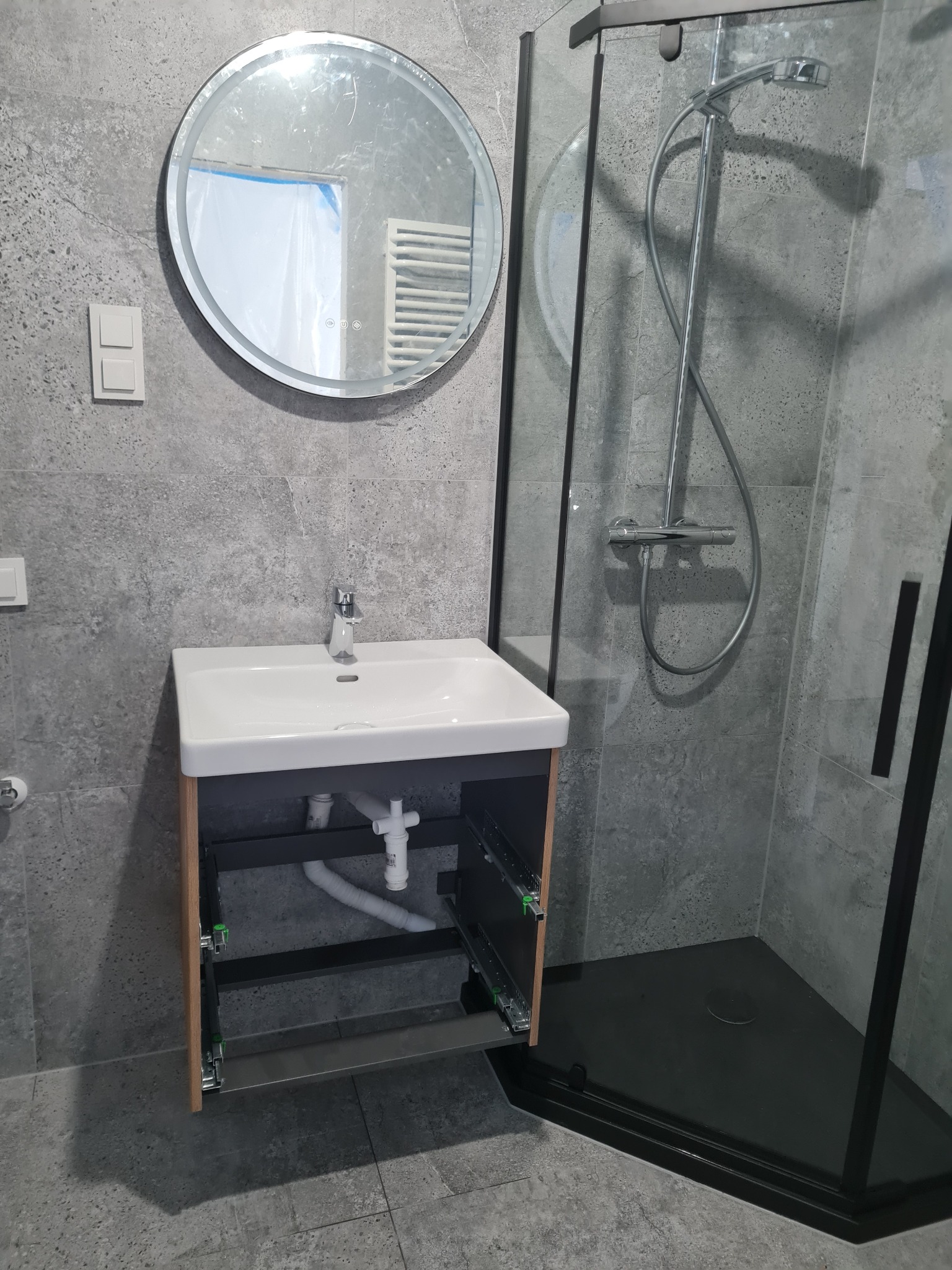 Szara łazienka z kabiną prysznicową typu walk-in z czarnymi profilami, okrągłym lustrem z podświetleniem i szafką pod umywalką z częściowo wysuniętą szufladą ukazującą metalowe prowadnice.