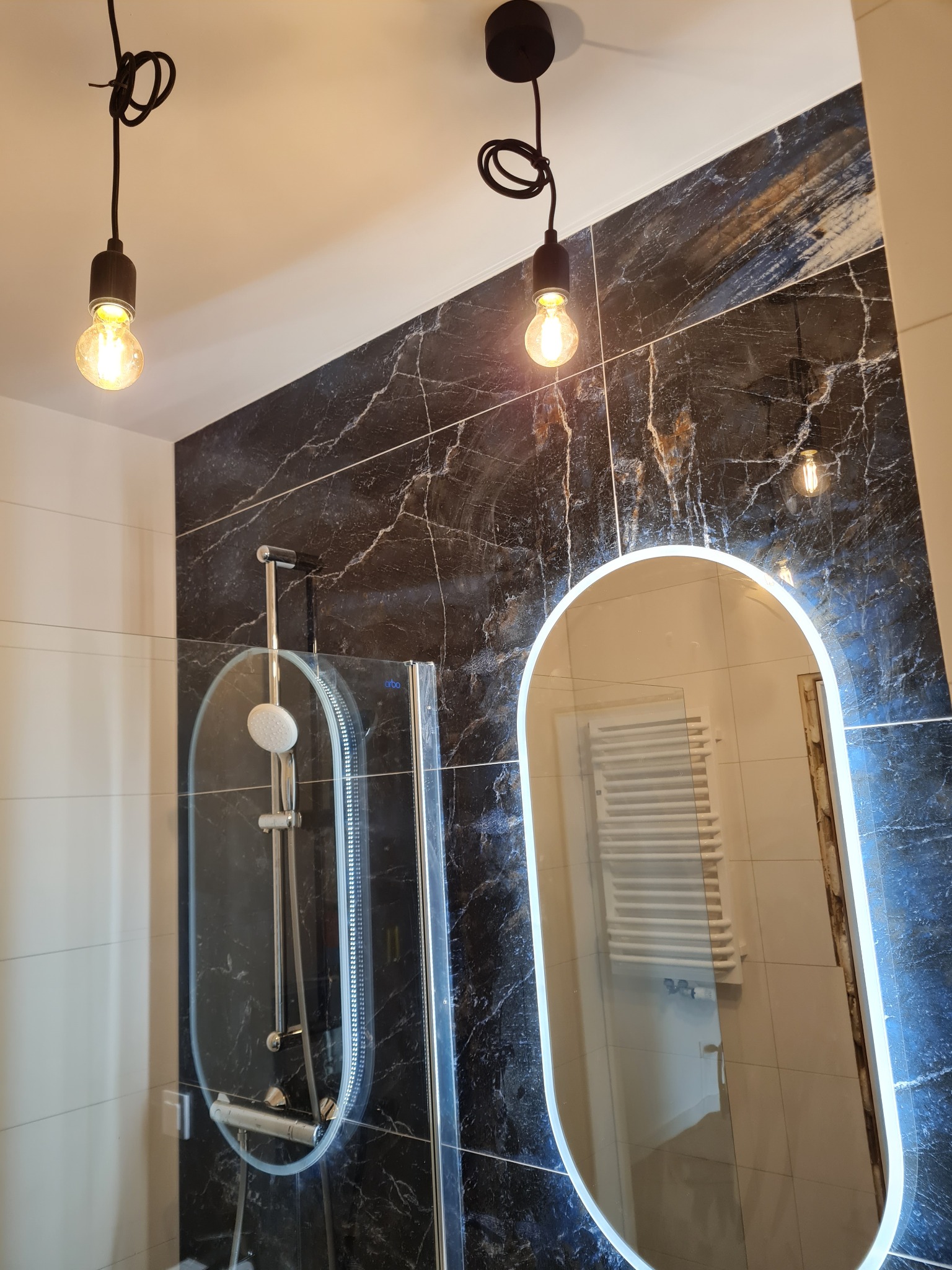 Nowoczesna łazienka z kabiną prysznicową i podświetlanym lustrem na tle płytek imitujących marmur. Dwie wiszące lampy z żarówkami Edisona.
