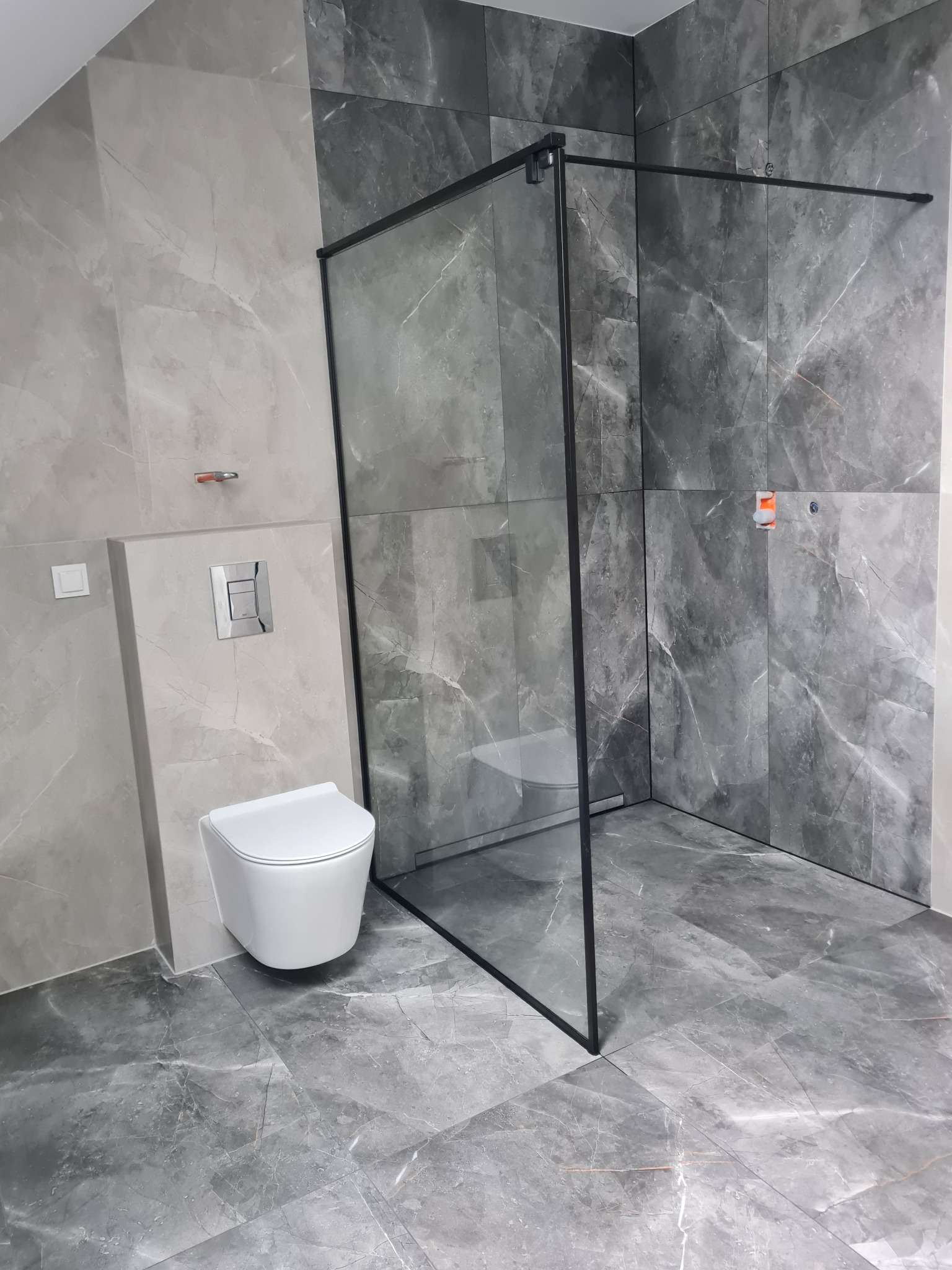 Szara łazienka z kabiną walk-in i podwieszaną toaletą. Płytki imitujące marmur na ścianach i podłodze. Minimalistyczny design, czarne ramy kabiny.