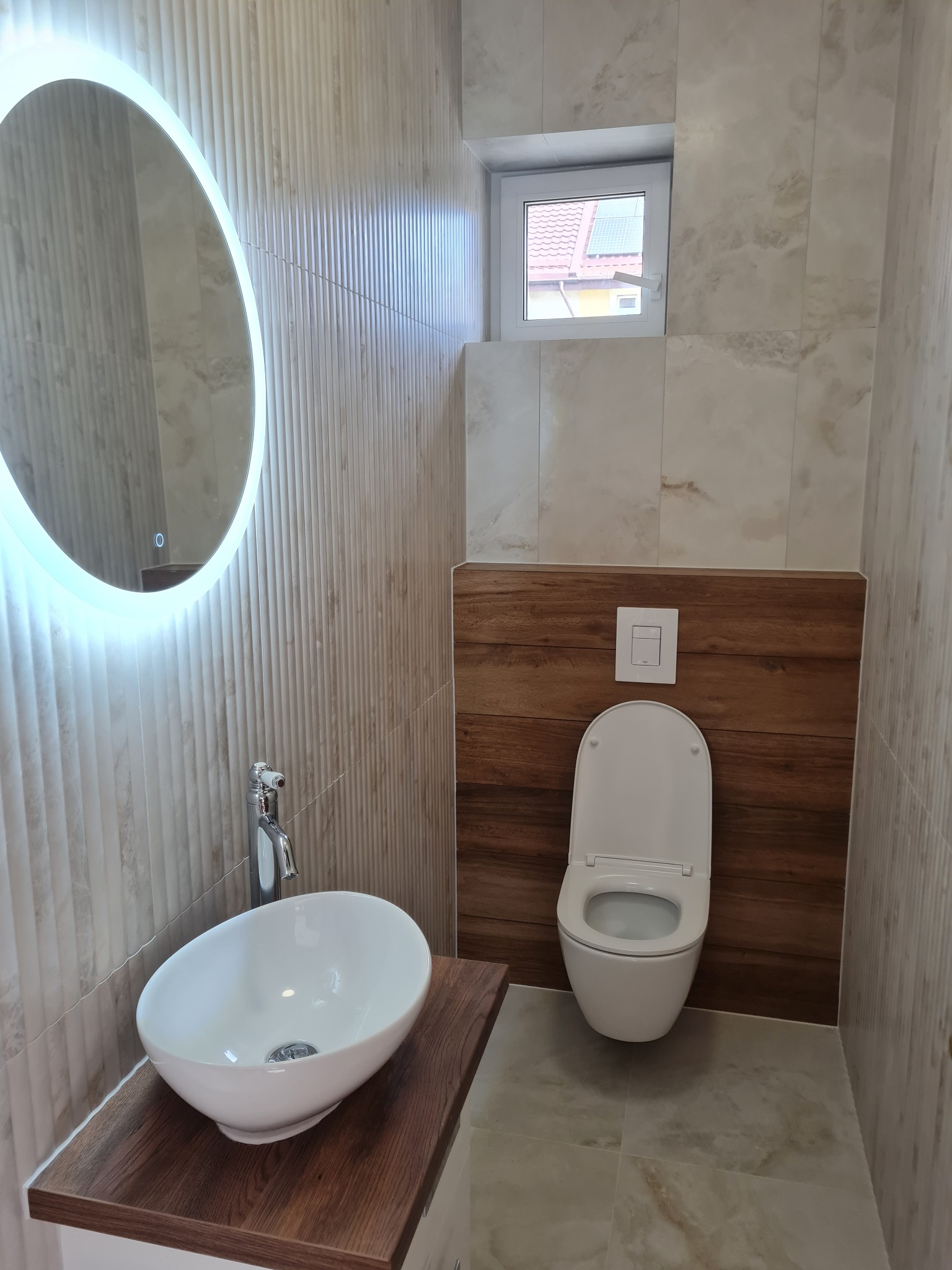Nowoczesna łazienka z owalną umywalką na drewnianym blacie, podświetlanym lustrem i toaletą na tle drewnopodobnych paneli. Jasne płytki na ścianach i podłodze.