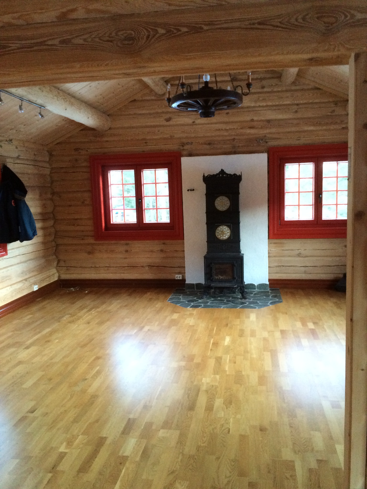 Drewniane wnętrze domu z parkietem, piecem kaflowym i czerwonymi ramami okiennymi. Widoczny fragment sufitu z drewnianych bali i nietypowy żyrandol.