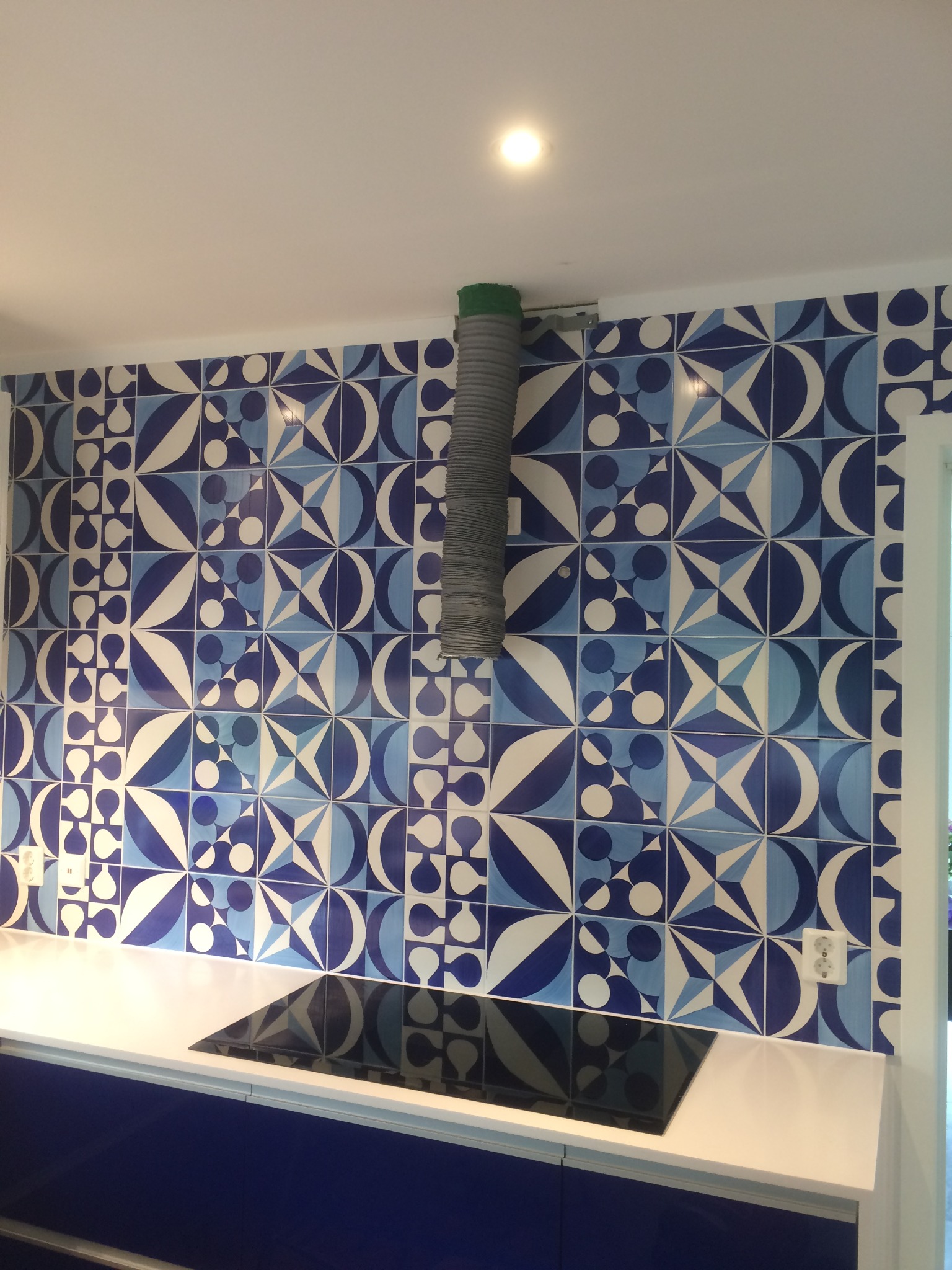 Kuchnia z niebiesko-białymi, wzorzystymi płytkami na ścianie, biały blat i niebieskie szafki, widoczny fragment okapu z szarą rurą.