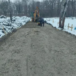 Przygotowanie podłoża pod posadzkę w Kątach Wrocławskich: pracownik zagęszcza ziemię ubijarką wibracyjną obok koparki, zimowy krajobraz z resztkami śniegu.