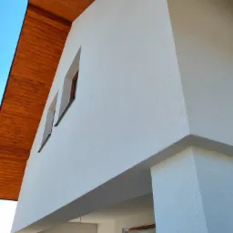 Fragment nowo wybudowanego domu z białą elewacją o fakturze baranka, drewnianym podbitką i dwoma wąskimi oknami, widok z dołu na tle błękitnego nieba.