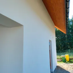 Fragment nowo otynkowanej, jasnej elewacji budynku z drewnianą podbitką i widocznymi rynnami, obok drzwi z zabezpieczeniem i śmietnikami w tle.