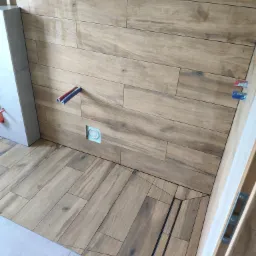 Ułożone płytki imitujące drewno na ścianie i podłodze w łazience, widoczny odpływ liniowy, wystające rurki instalacji wodnej i otwór na gniazdko elektryczne.