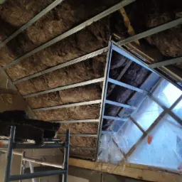 Montaż izolacji z wełny mineralnej na poddaszu, widoczne metalowe profile, rusztowanie i folia ochronna na oknie dachowym.