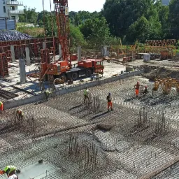 Szeroki plan budowy fundamentów z widocznym zbrojeniem, pracownikami w odblaskowych kamizelkach, żurawiem i częściowo wylanym betonem na tle budynku mieszkalnego i zieleni.