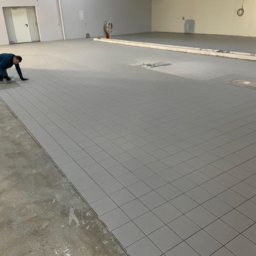 PMP remonty - Układanie szarych płytek podłogowych w dużym pomieszczeniu; pracownik w niebieskiej bluzie i ciemnych spodniach klęczy przy krawędzi ułożonych płytek, obok wózek z narzędziami, widoczna surowa...