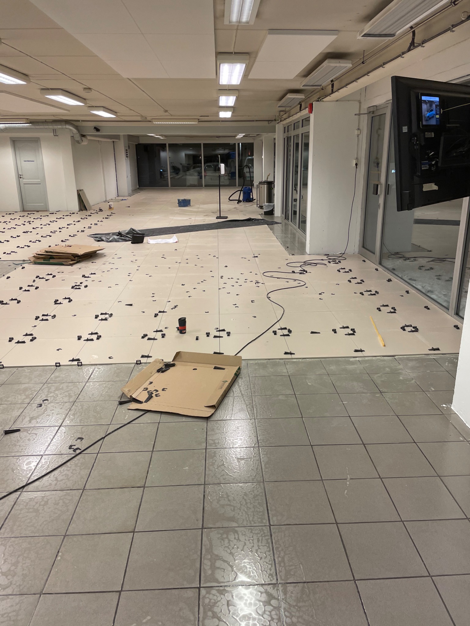 Wnętrze w trakcie układania jasnych płytek podłogowych z użyciem czarnych klipsów poziomujących, widoczne kartony, poziomica i kabel zasilający na podłodze.