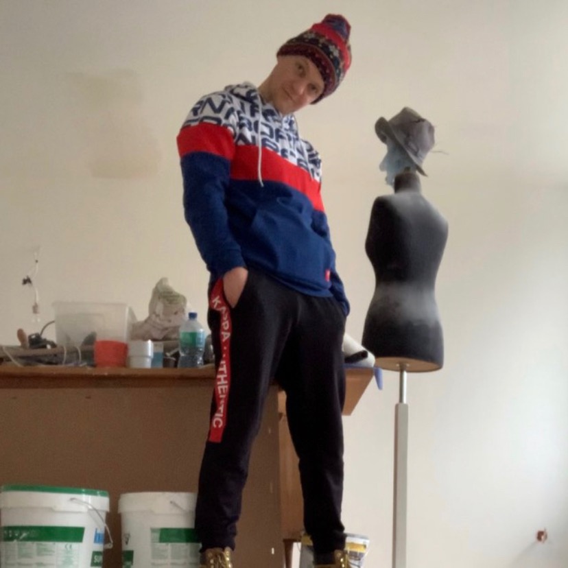 Młody mężczyzna w czapce i bluzie stoi na tle pokoju w trakcie prac, widoczne wiadra z farbą i manekin z kapeluszem.