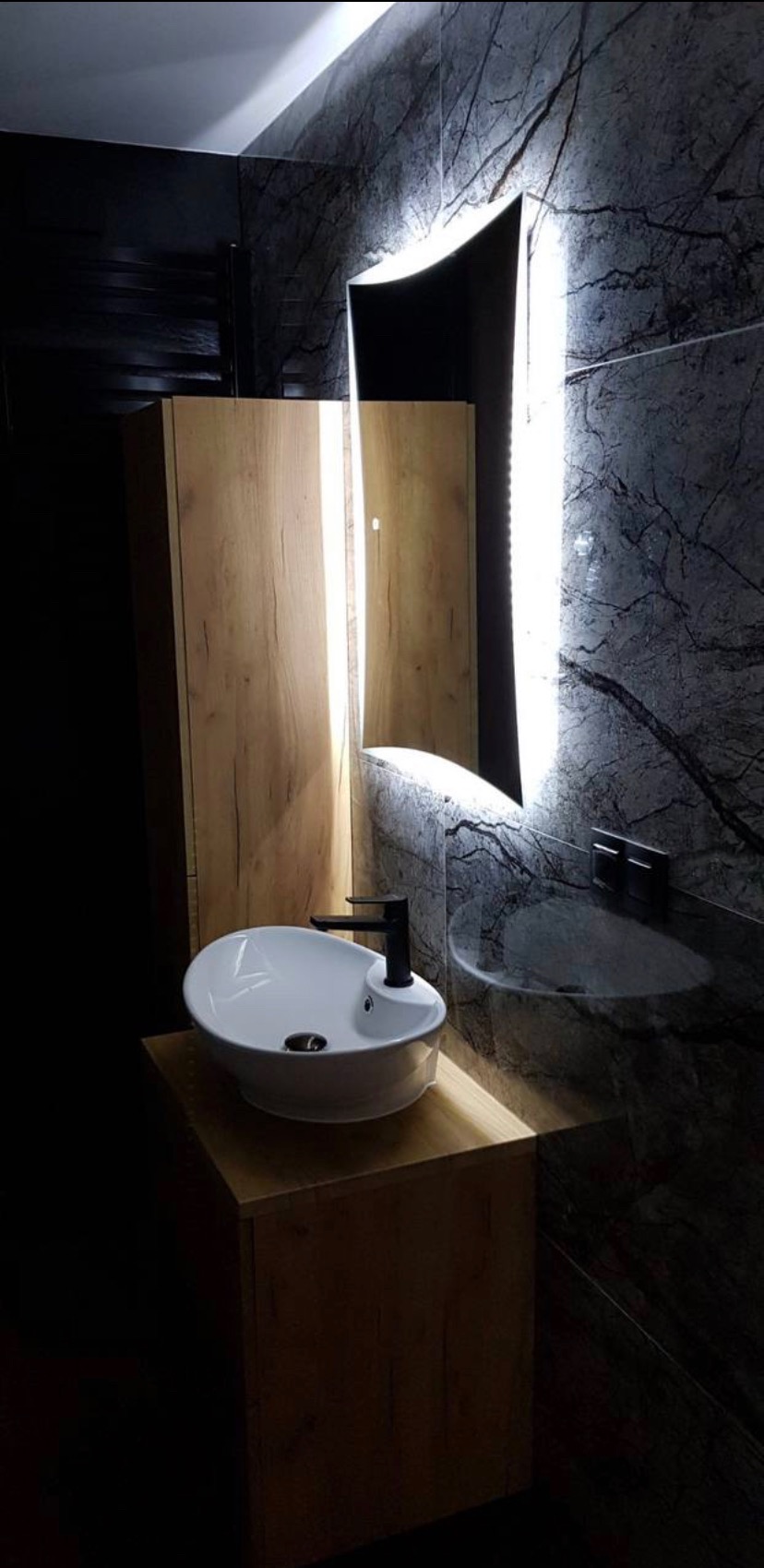 Nowoczesna łazienka z umywalką nablatową o owalnym kształcie i czarną armaturą, podświetlane lustro o nieregularnym kształcie, ściana wykończona płytkami imitującymi ciemny marmur z wyraźnym...
