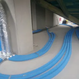 Montaż rekuperacji w surowym wnętrzu: Niebieskie rury wentylacyjne prowadzone po podłodze, połączone srebrną, izolowaną rurą do rozdzielacza, widoczne elementy konstrukcji dachu i agregat w tle.