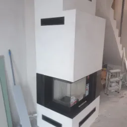 Nowoczesny, biały kominek z czarnymi akcentami w trakcie montażu, widoczne elementy instalacji i narzędzia, surowe wnętrze w trakcie wykańczania.