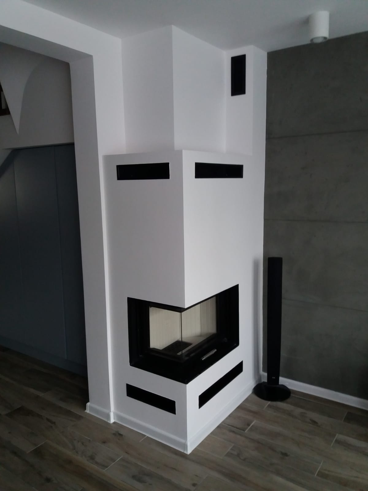 Nowoczesny, narożny kominek z białym obramowaniem i czarnymi akcentami wentylacyjnymi, umieszczony w rogu pomieszczenia z szarą ścianą i drewnopodobną podłogą.