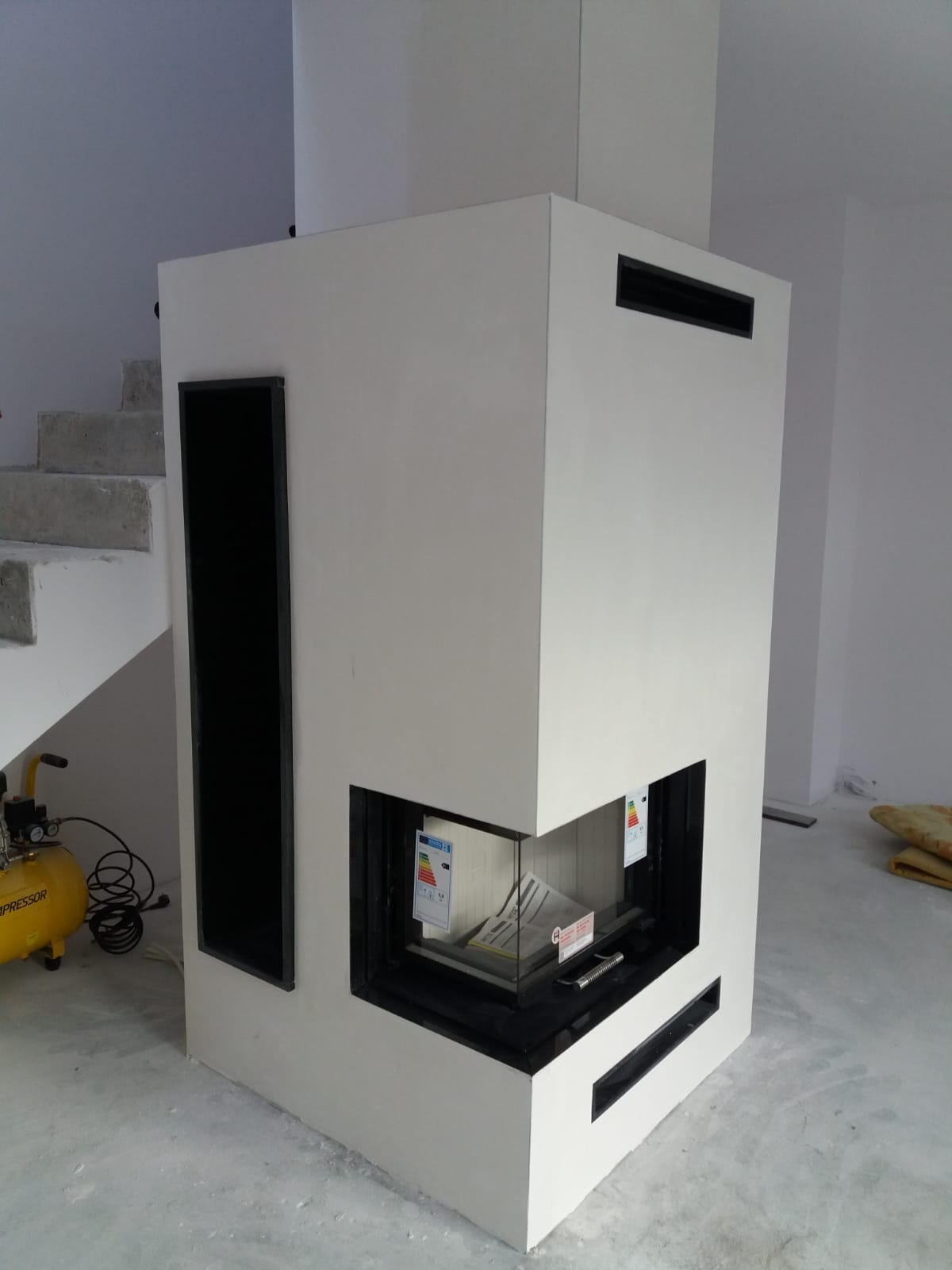 Nowoczesny, biały kominek narożny z czarnymi akcentami, widoczne schody w tle i żółty kompresor, pomieszczenie w trakcie wykańczania.