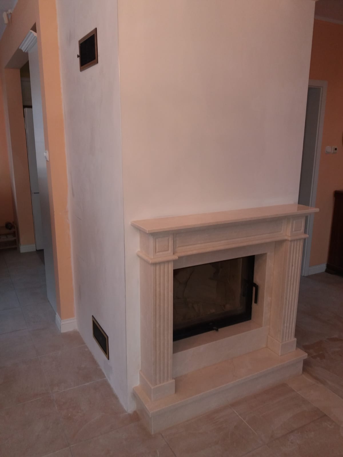 Klasyczny kominek z obudową z jasnego kamienia, widoczny fragment ściany z kratką wentylacyjną, beżowe płytki podłogowe.