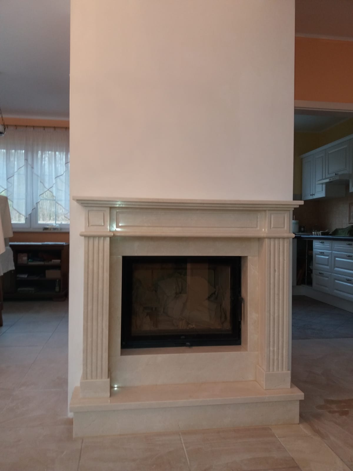 Elegancki kominek z obudową z jasnego kamienia, widoczny w salonie z elementami kuchni w tle.