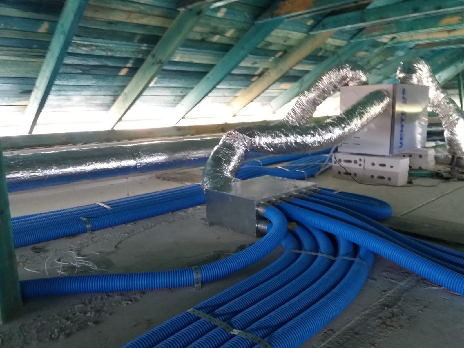 Instalacja systemów wentylacyjnych: rozdzielacz z podłączonymi niebieskimi rurami wentylacyjnymi w surowym stanie poddasza, widoczna izolacja i elementy mocujące.