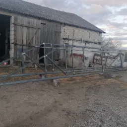 Stalowa konstrukcja szkieletowa, prawdopodobnie brama, oparta na kamieniach przed starym budynkiem gospodarczym z cegły, widok z zewnątrz.