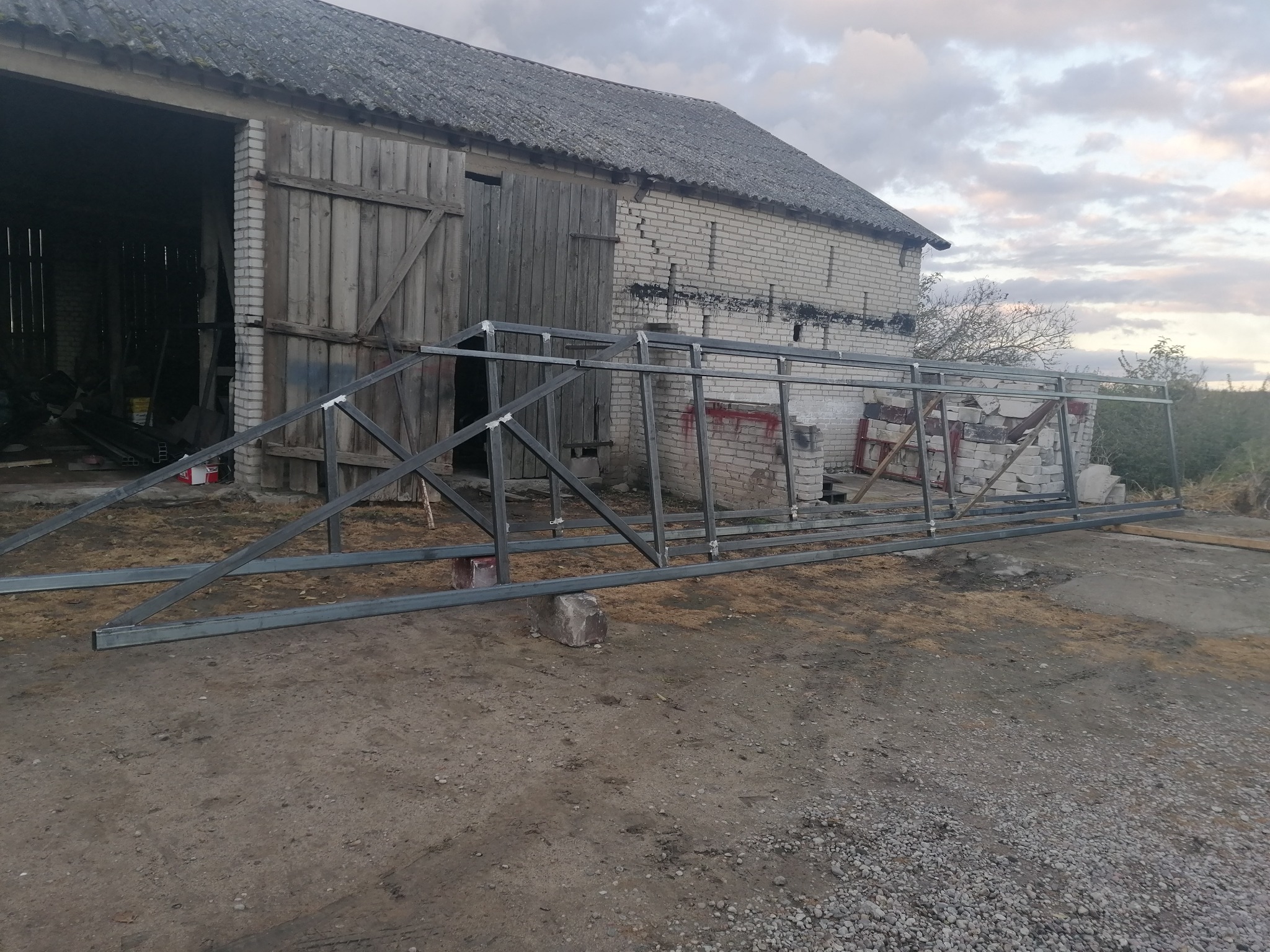 Stalowa konstrukcja szkieletowa, prawdopodobnie brama, oparta na kamieniach przed starym budynkiem gospodarczym z cegły, widok z zewnątrz.