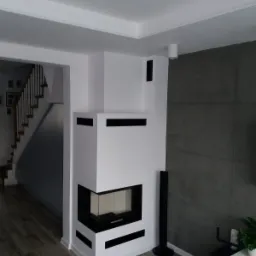 Nowoczesny, biały kominek narożny z czarnymi akcentami w salonie, widoczne schody w tle, szara ściana z płytek imitujących beton.