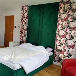 Sypialnia z ł&oacute;żkiem o zielonej ramie i wezgłowiu z paneli tapicerowanych, ściana z tapetą w kwiaty, czerwony fotel w rogu, widok na drzwi i zapakowane meble.