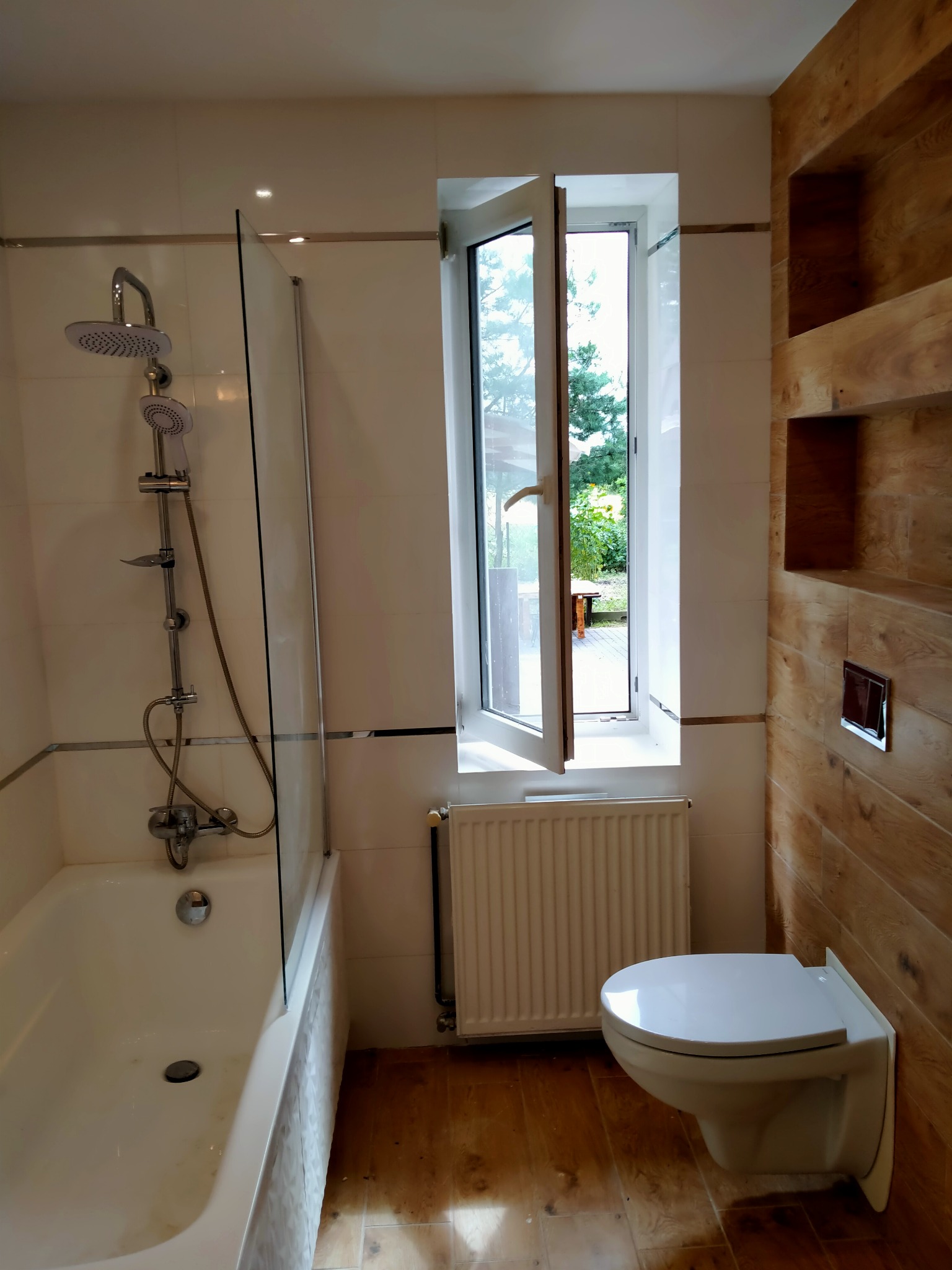 Wykończona łazienka z wanną z parawanem, toaletą podwieszaną i drewnopodobnymi płytkami na ścianie z wnękami, widok na otwarte okno z zielenią na zewnątrz.