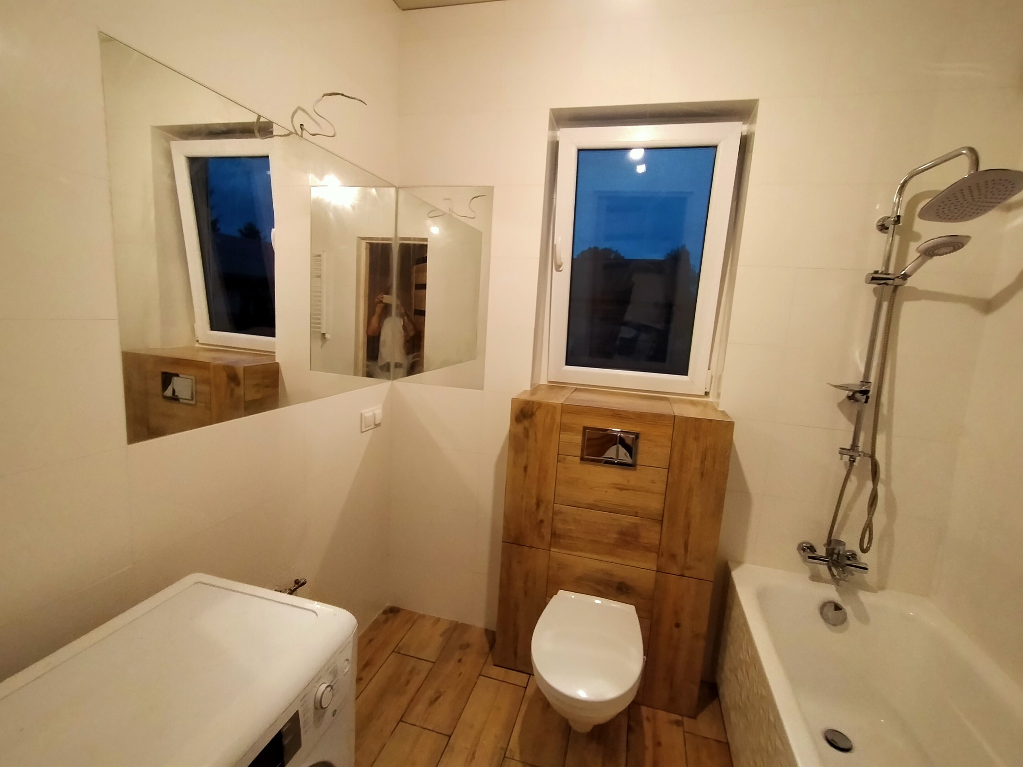 Wykończona łazienka z drewnianymi akcentami: obudowa toalety, drewniana podłoga, biała wanna, pralka i lustro na ścianie.